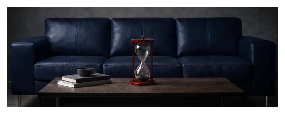 Salón oscuro con un sofa negro al fondo y un reloj de arena delante